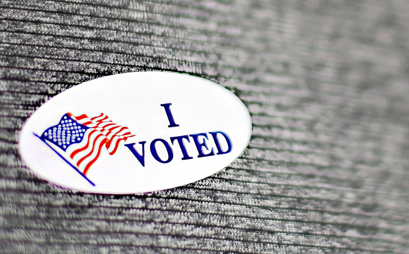 Una pegatina ovalada blanca con las palabras "I VOTED" en letras azules y una bandera estadounidense ondeante en el lado izquierdo, sobre un fondo texturizado gris oscuro. Foto: Imagen de Pixabay.