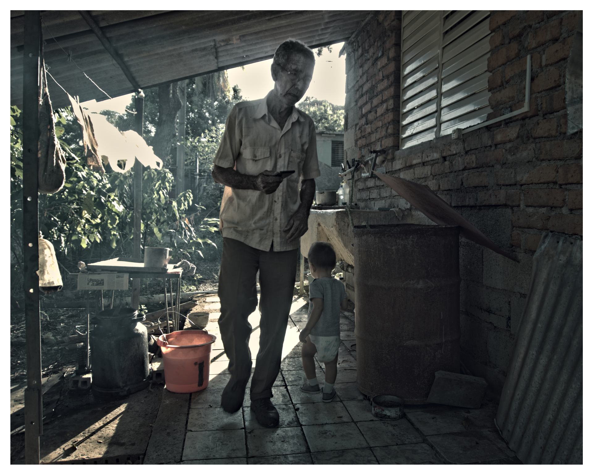  Hombre mayor caminando bajo un techo improvisado junto a un niño pequeño, en un área de lavado exterior con paredes de ladrillo.