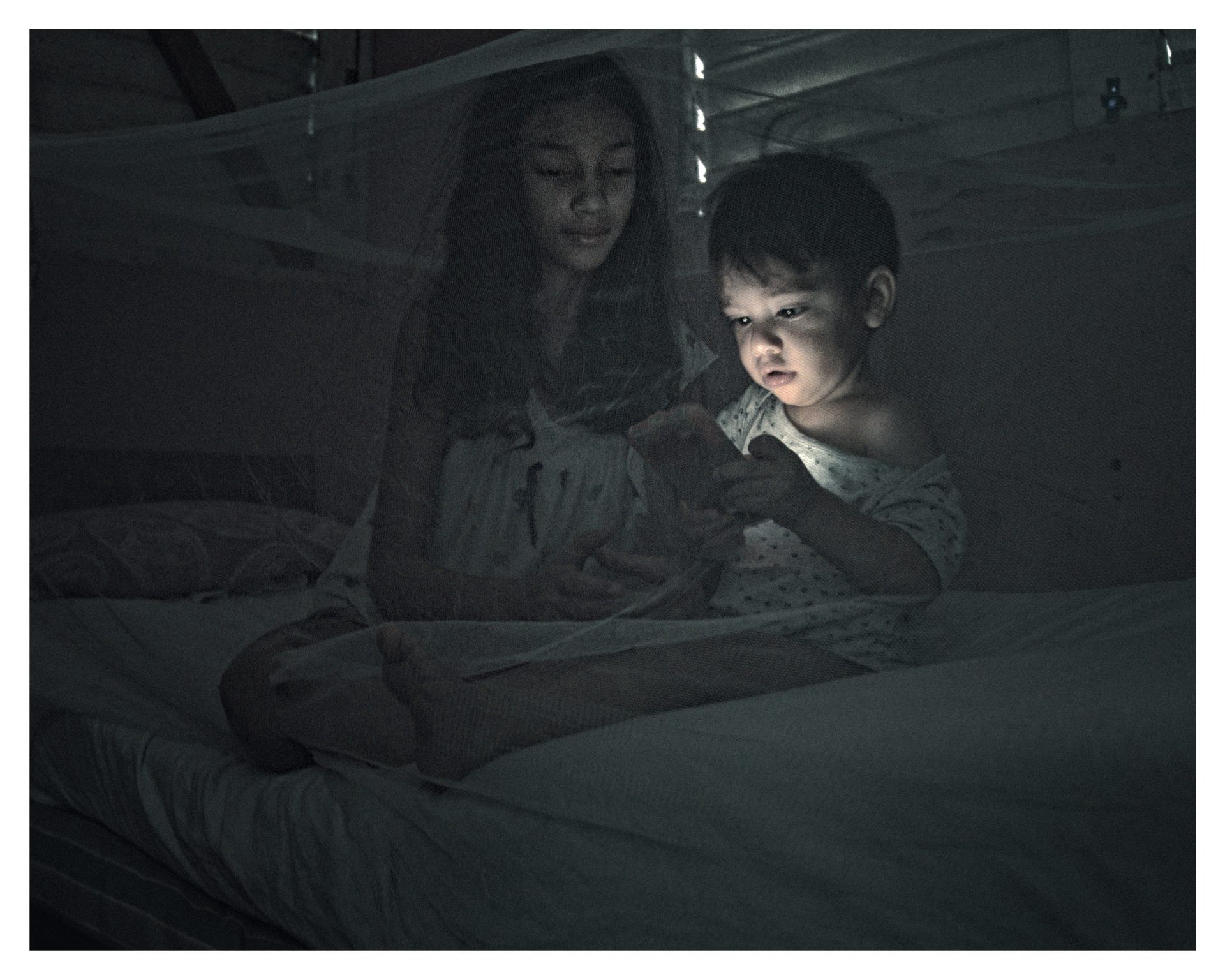 Niño pequeño y una niña sentados en la cama bajo un mosquitero, iluminados por la luz de un celular.