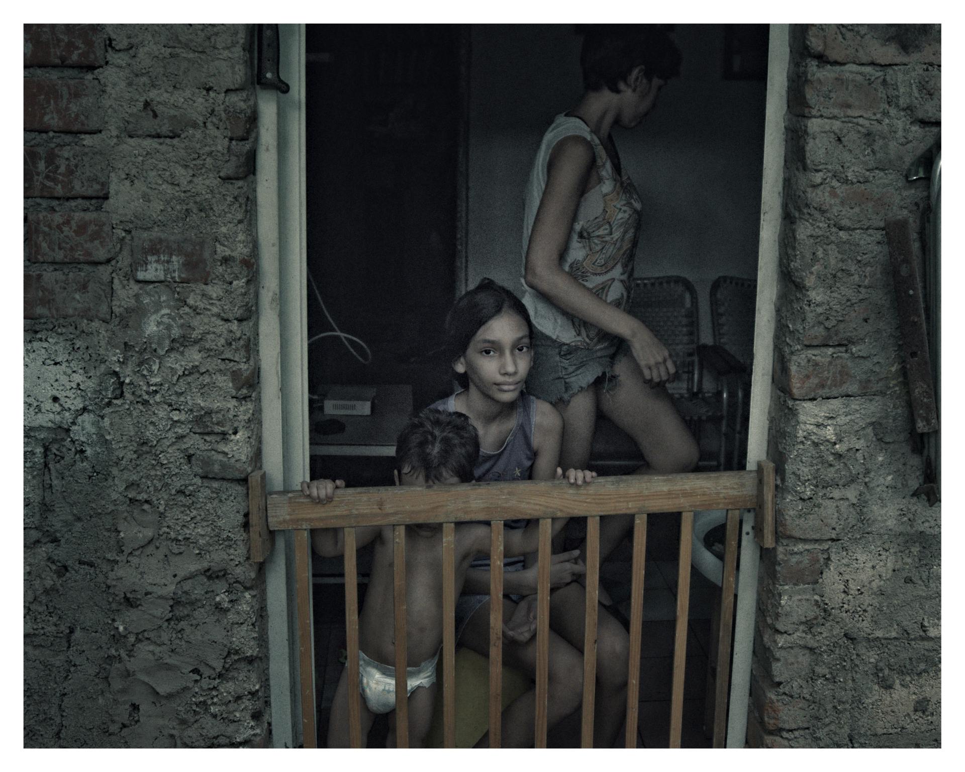 Tres personas en la entrada de una casa de ladrillo sin repellar; una niña mira al frente mientras un niño pequeño se sostiene de una baranda y una mujer camina detrás.