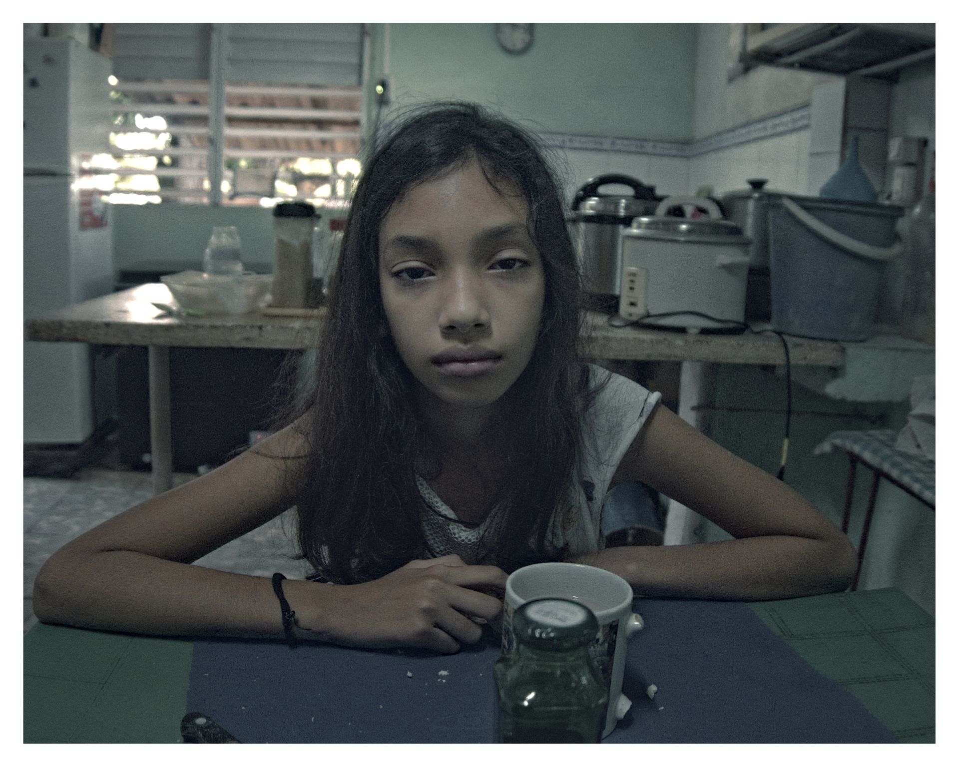 Niña con expresión seria sentada en una cocina modesta, con tazas y objetos domésticos sobre la mesa.