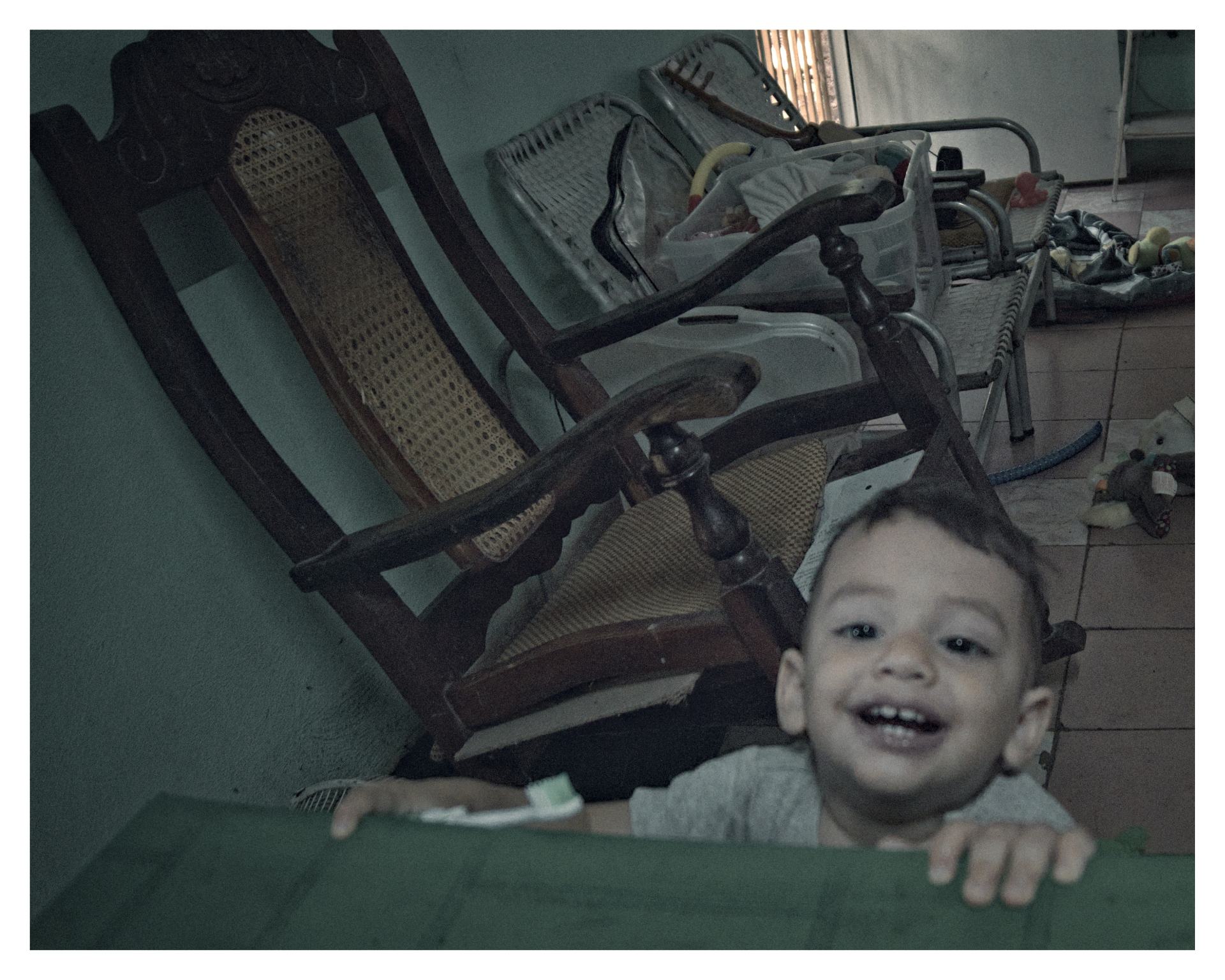 Niño pequeño sonriente asomado a una mesa, en una sala con muebles antiguos y juguetes dispersos.