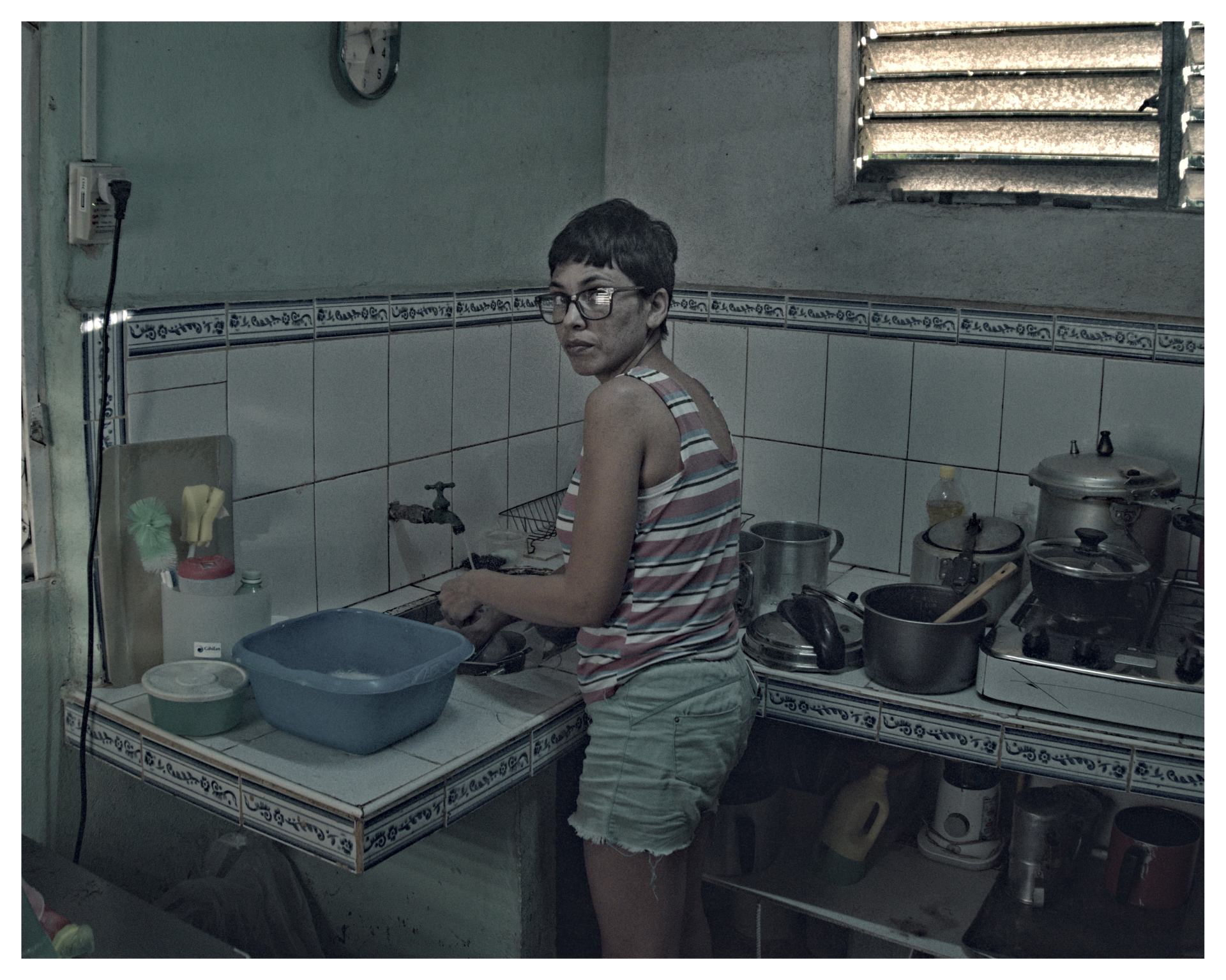 Mujer lavando en una cocina modesta, rodeada de ollas y utensilios, mientras mira hacia la cámara.