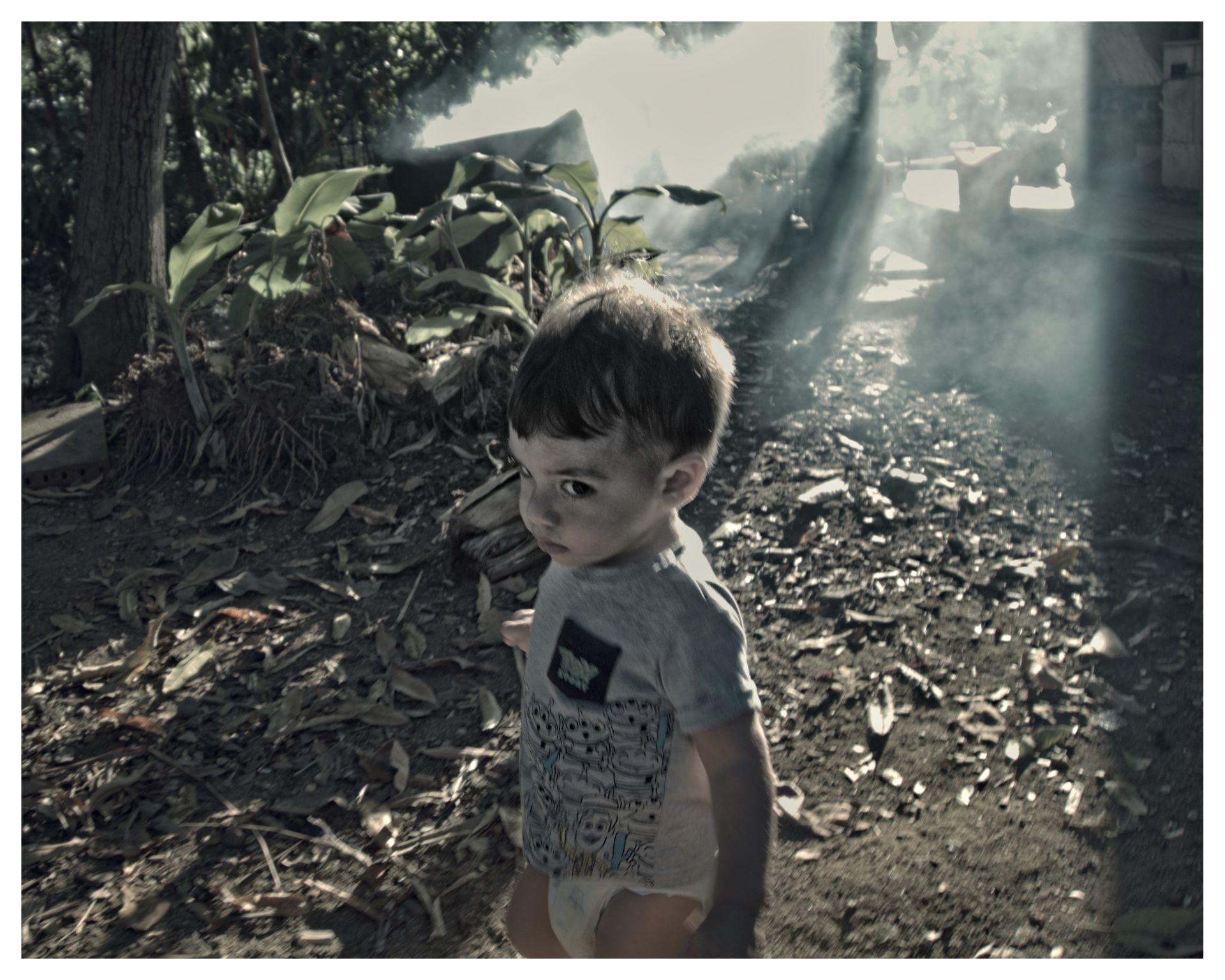 Niño pequeño caminando entre hojas secas en un patio sombreado, con rayos de luz y humo en el fondo.