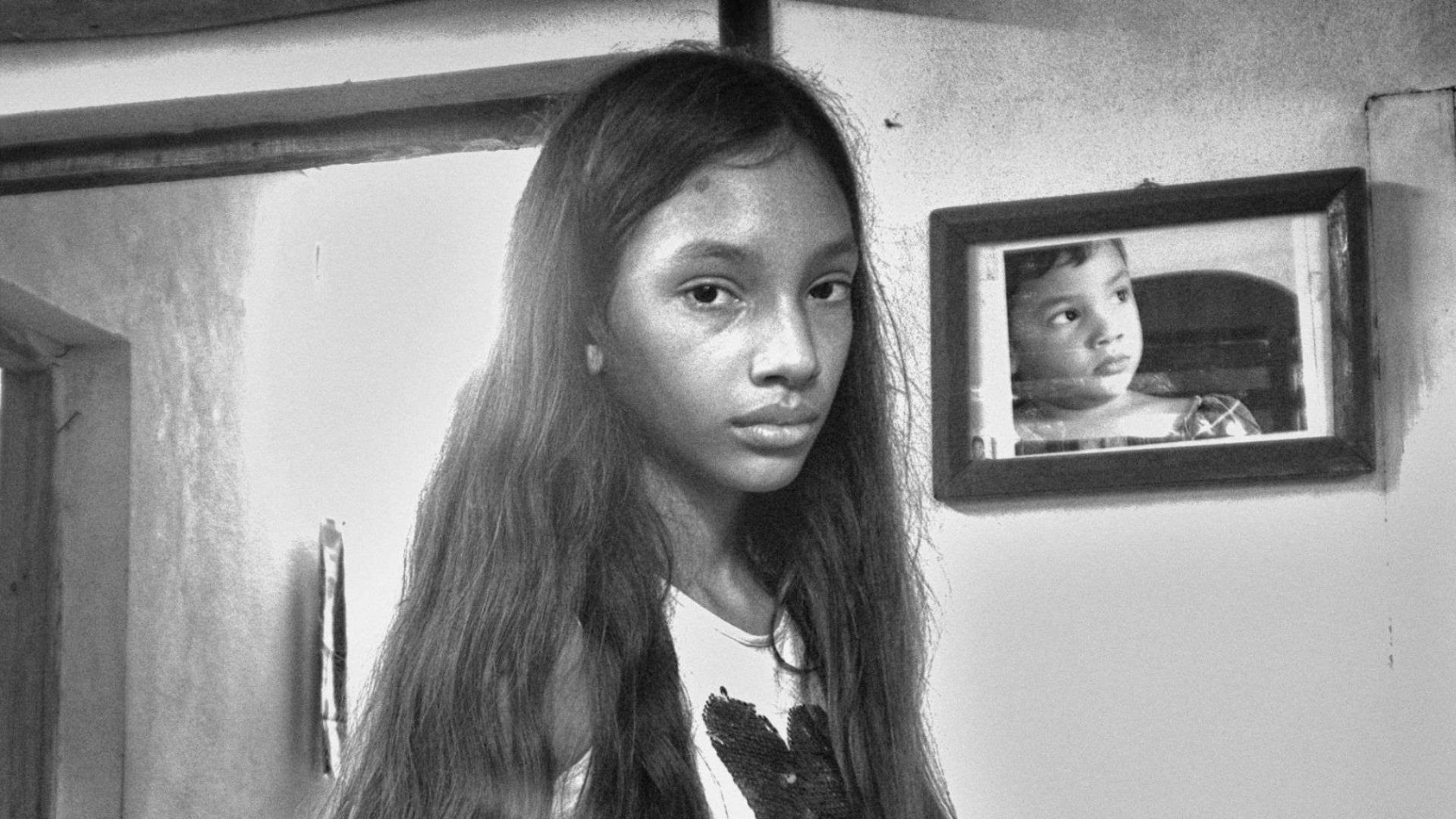 Niña con expresión seria en el interior de una casa modesta, con un retrato infantil en la pared.