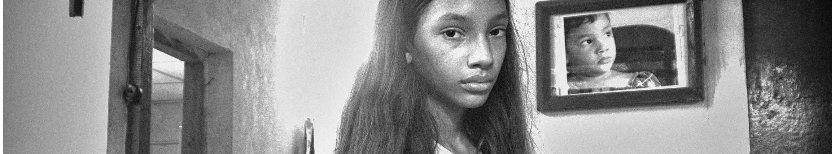 Niña con expresión seria en el interior de una casa modesta, con un retrato infantil en la pared.