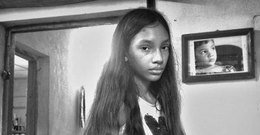 Niña con expresión seria en el interior de una casa modesta, con un retrato infantil en la pared.