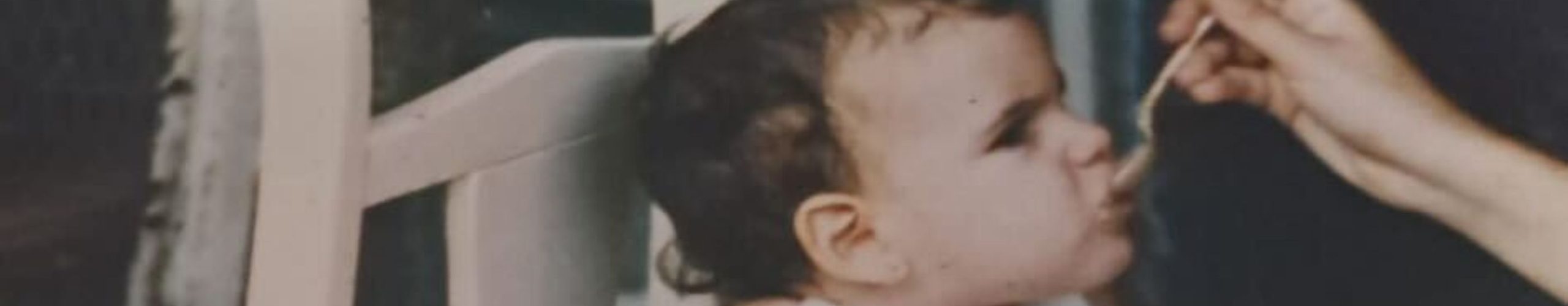 Bebé sentada en una silla alta de madera mientras una mano adulta le da de comer con una cucharita.