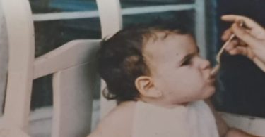 Bebé sentada en una silla alta de madera mientras una mano adulta le da de comer con una cucharita.