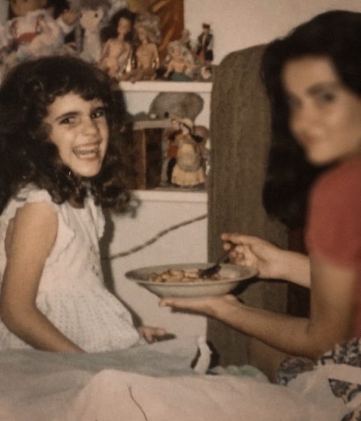 Niña sonriente junto a su madre mientras esta le ofrece un plato de comida, sentadas sobre una cama en Cuba.
