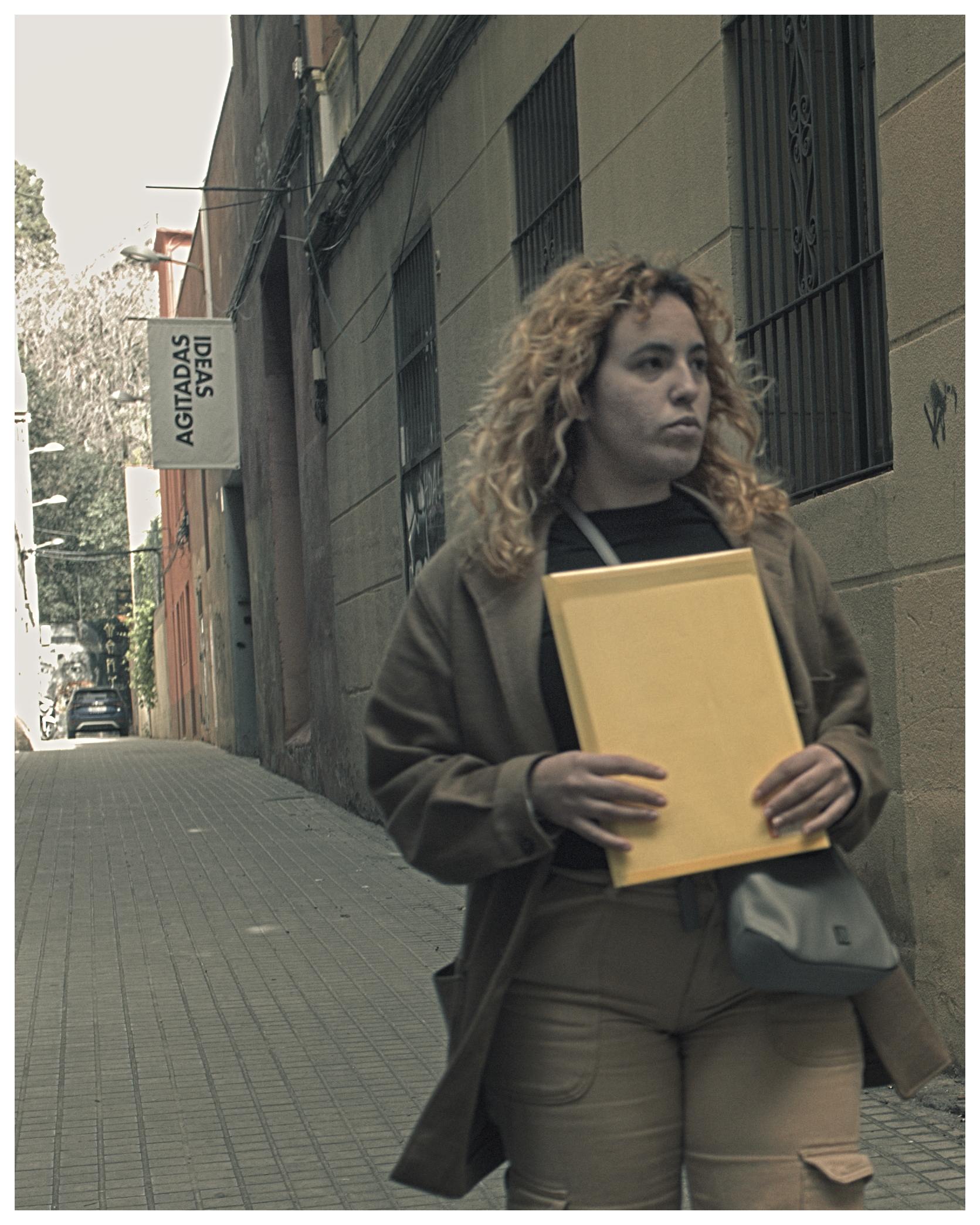 Mujer migrante camina por una calle con gesto serio, cargando sobres con documentos.