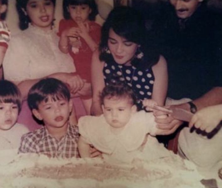 Niña pequeña frente a un pastel de cumpleaños, rodeada de niños y adultos durante una celebración.