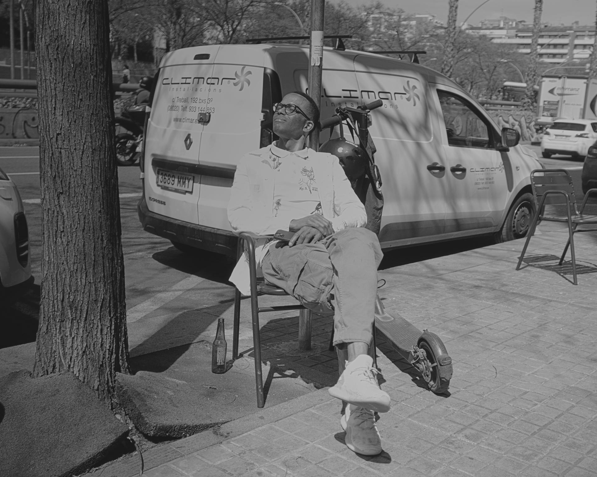 Hombre sentado junto a una scooter, tomando el sol en la acera.