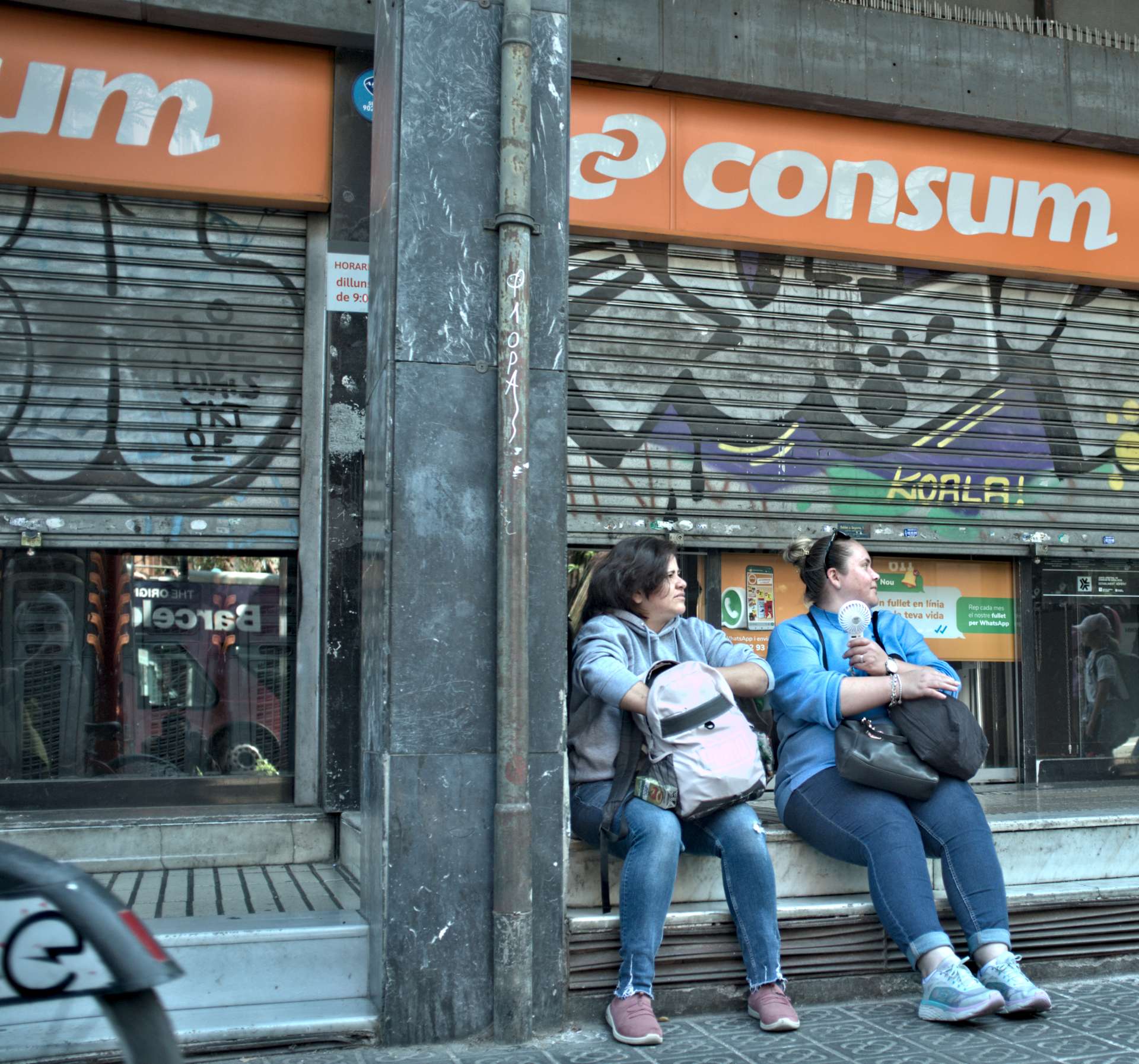 Dos mujeres se abanican sentadas frente a una tienda cerrada.