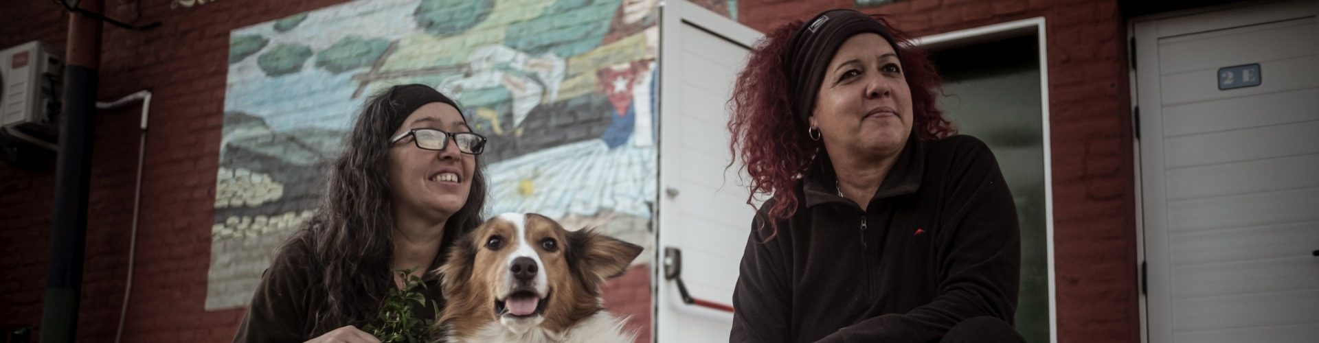 Geraidy Brito Montes de Oca y Yaqui Saiz, junto a su perro, sentadas frente al mural de la Casa de Títeres Cubana Argentina en Junín. Detrás, se ve la fachada del espacio cultural con puertas blancas y paredes de ladrillo rojo. Foto: Ella Fernández.