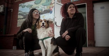 Geraidy Brito Montes de Oca y Yaqui Saiz, junto a su perro, sentadas frente al mural de la Casa de Títeres Cubana Argentina en Junín. Detrás, se ve la fachada del espacio cultural con puertas blancas y paredes de ladrillo rojo. Foto: Ella Fernández.