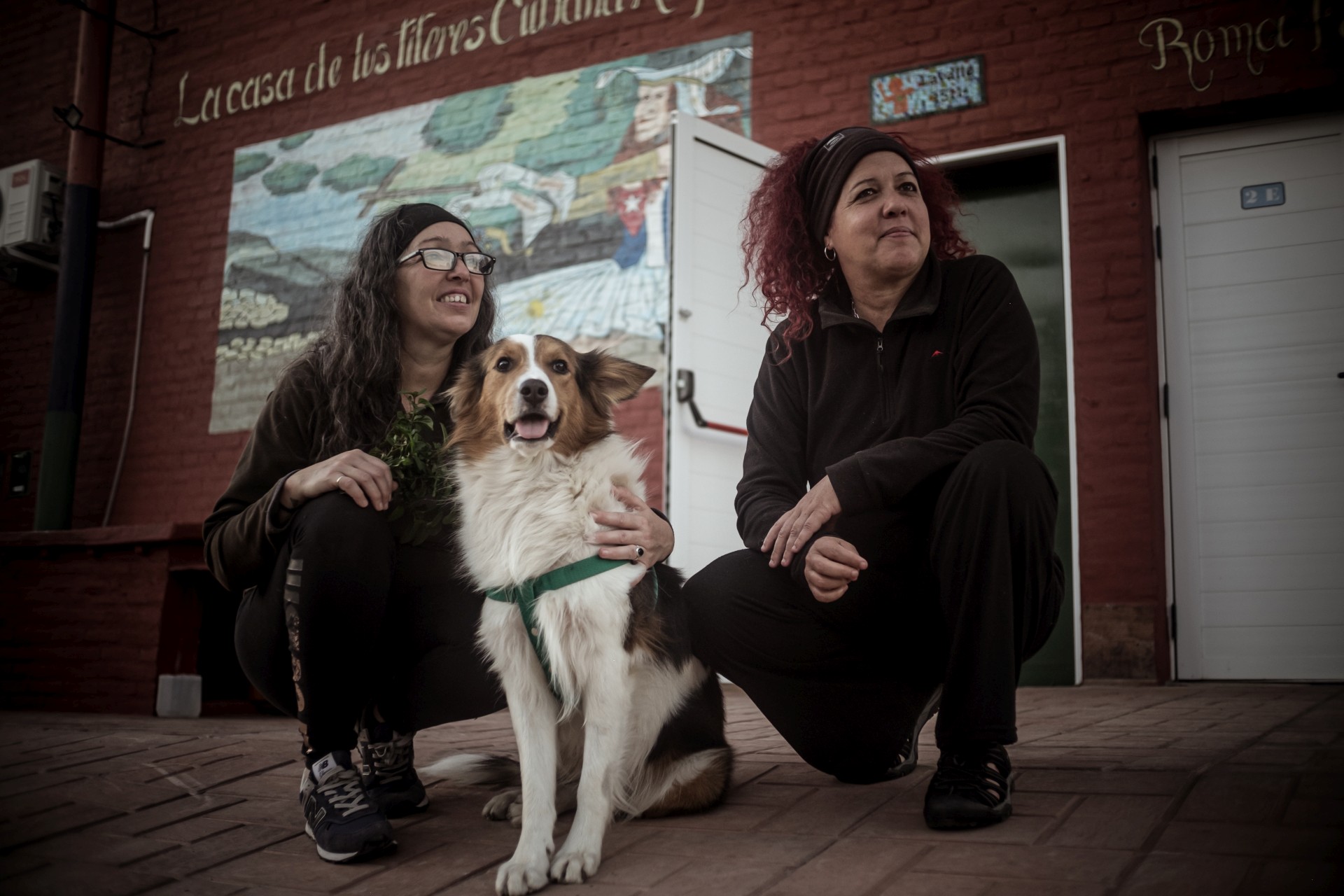 Geraidy Brito Montes de Oca y Yaqui Saiz, junto a su perro, sentadas frente al mural de la Casa de Títeres Cubana Argentina en Junín. Detrás, se ve la fachada del espacio cultural con puertas blancas y paredes de ladrillo rojo. Foto: Ella Fernández.