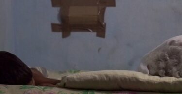 Hacinamiento en Santiago de Cuba. Dos personas (un niño y una adulta mayor) duermen en una cama estrecha en un cuarto pequeño y oscuro, simbolizando el agotamiento de una madre soltera en Cuba.