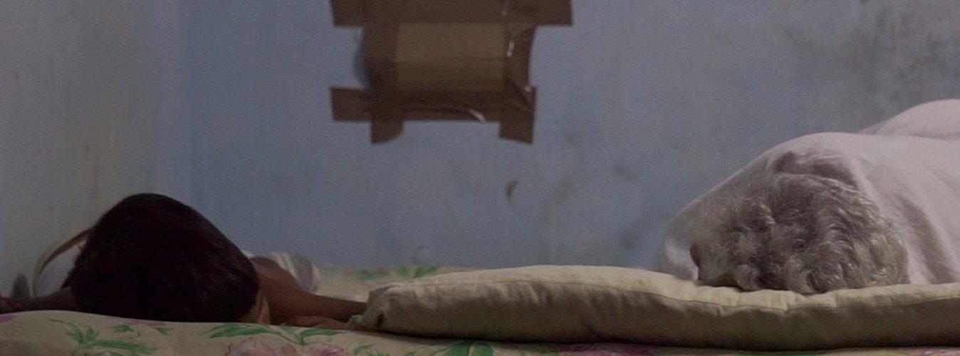 Hacinamiento en Santiago de Cuba. Dos personas (un niño y una adulta mayor) duermen en una cama estrecha en un cuarto pequeño y oscuro, simbolizando el agotamiento de una madre soltera en Cuba.